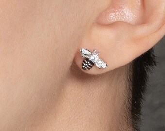 May include: A close-up of a person's ear with a small silver bee stud earring. The bee has black and white details.