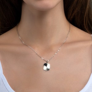 May include: A woman with long brown hair is wearing a white vest top and a silver necklace with a small pendant. She is looking at the camera and smiling.