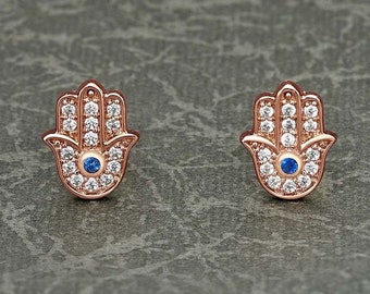 May include: A pair of rose gold earrings featuring a hamsa hand design with a blue evil eye in the centre. Each earring is adorned with clear cubic zirconia stones.