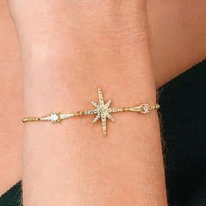 May include: A gold bracelet with a starburst charm and small clear stones. The bracelet is on a woman's wrist.