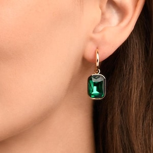 May include: A woman's ear adorned with a gold hoop earring featuring a rectangular, emerald-green gemstone. The earring's design is simple, highlighting the gemstone's vivid green hue. The woman has fair skin and brown hair.