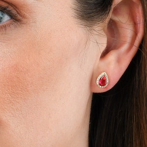 Könnte beinhalten: Eine Frau trägt ein Paar kleine, tropfenförmige Ohrringe mit roten Edelsteinen und einer goldenen Fassung. Die Ohrringe sind vor einem blauen Hintergrund platziert.