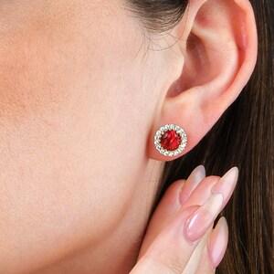 May include: A woman with long brown hair wearing a red gemstone earring with a clear crystal halo.