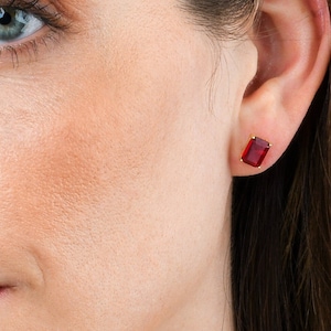 Pode incluir: Um close-up do rosto de uma mulher com brincos de pedras preciosas vermelhas. A mulher está usando um colar de ouro e tem o cabelo puxado para trás.