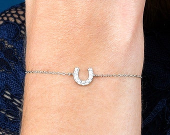 Puede incluir: Una pulsera de plata con un dije de herradura adornado con pequeñas piedras brillantes. La pulsera es delicada y se muestra en una muñeca sobre un fondo azul. El dije de herradura es el punto focal de la joya.