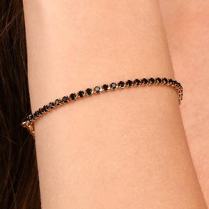 Puede incluir: Una delicada pulsera de tenis que presenta una hilera continua de pequeñas piedras negras redondas y facetadas engastadas en una fina banda metálica. La pulsera rodea la muñeca, mostrando un diseño uniforme con un cierre visible en el lateral.