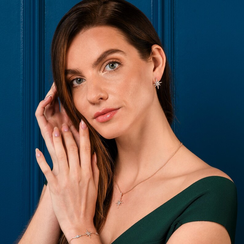 May include: A woman wearing a matching jewelry set featuring starburst designs. The set includes a necklace, earrings, and bracelet, all in a rose gold tone. The woman is wearing a dark green dress against a blue background.