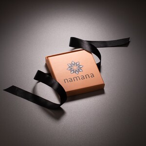 May include: A square, open, copper-colored box with the word "namana" printed on the front, along with a geometric design. A black ribbon is draped around the box. The box is on a dark gray surface.