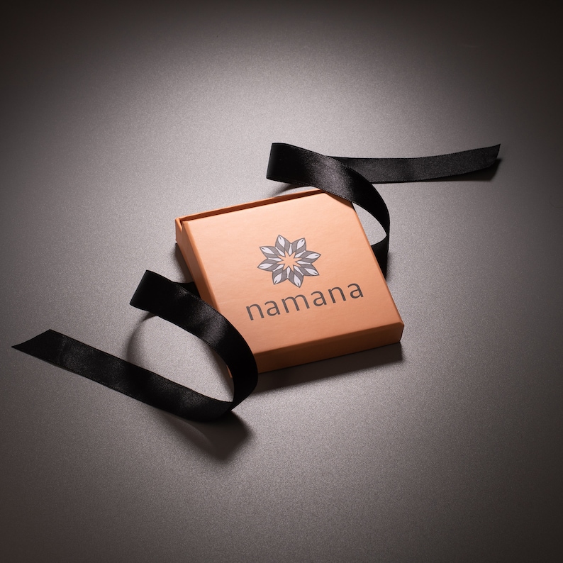 May include: A square, open, copper-colored box with the word "namana" printed on the front, along with a geometric design. A black ribbon is draped around the box. The box is on a dark gray surface.