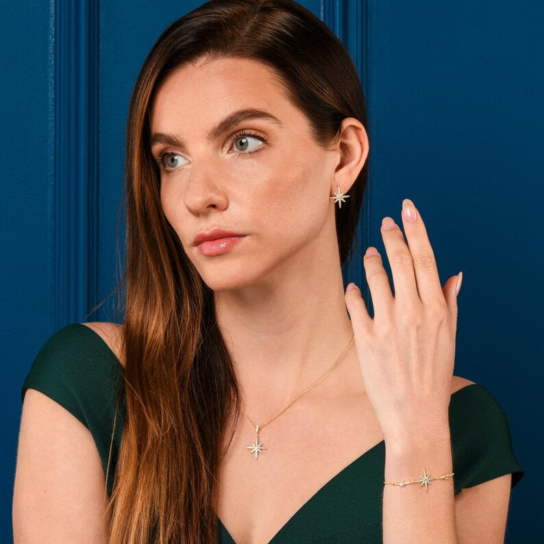 May include: A woman wearing a green dress with a gold necklace and earrings featuring a starburst design. The necklace and earrings are accented with small clear stones.