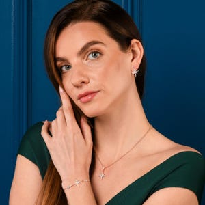 May include: A woman wearing a matching jewelry set featuring starburst designs. The set includes a necklace, earrings, and a bracelet. The jewelry is gold-toned and adorned with small, sparkling accents. The woman is wearing a dark green dress.