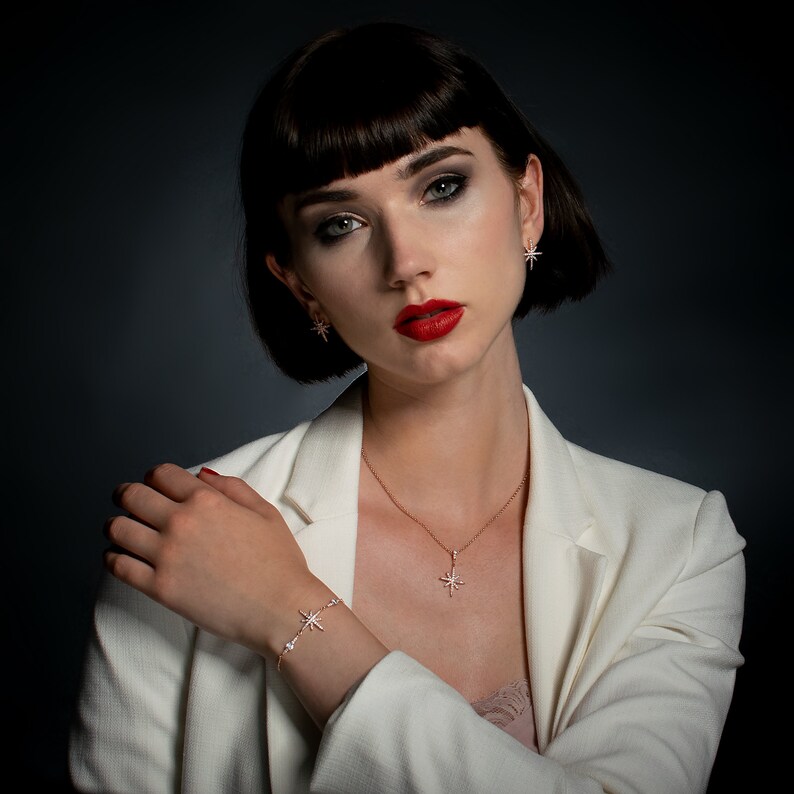 May include: A woman wearing a white blazer and a matching jewelry set. The jewelry includes a starburst pendant necklace, earrings, and a bracelet. The jewelry is silver-toned and features a star design. The woman has dark hair and red lipstick.