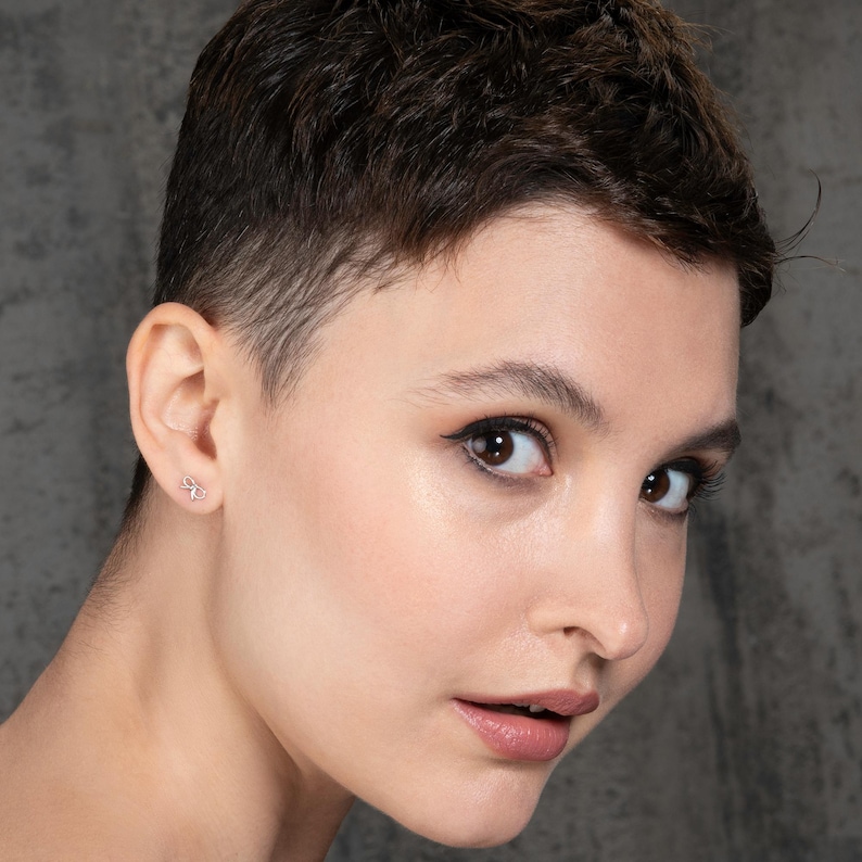 K&ouml;nnte beinhalten: Nahaufnahme eines Gesichts mit kurzen, dunklen Haaren. Die Person tr&auml;gt einen kleinen, silbernen Ohrring in Form einer Schleife.