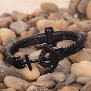 May include: A black anchor bracelet with a mesh design. The bracelet features a nautical anchor charm and a magnetic clasp. The bracelet is displayed on a bed of small, smooth stones.