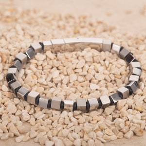 May include: A silver bracelet with black accents made of square-shaped beads. The bracelet is made of metal and has a clasp closure.