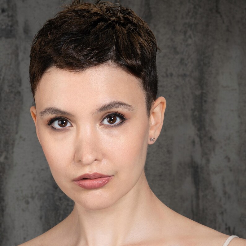 K&ouml;nnte beinhalten: Eine Frau mit kurzen, dunkelbraunen Haaren und einem neutralen Gesichtsausdruck. Sie tr&auml;gt ein schlichtes, helles Oberteil und hat ein dezentes Make-up.