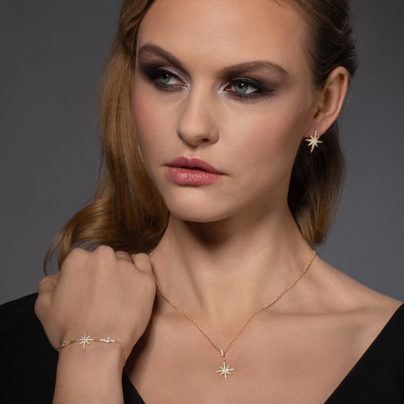 May include: A gold necklace, bracelet, and earrings featuring a starburst design with clear cubic zirconia stones. The jewelry is worn by a model with long blonde hair and dark eye makeup.