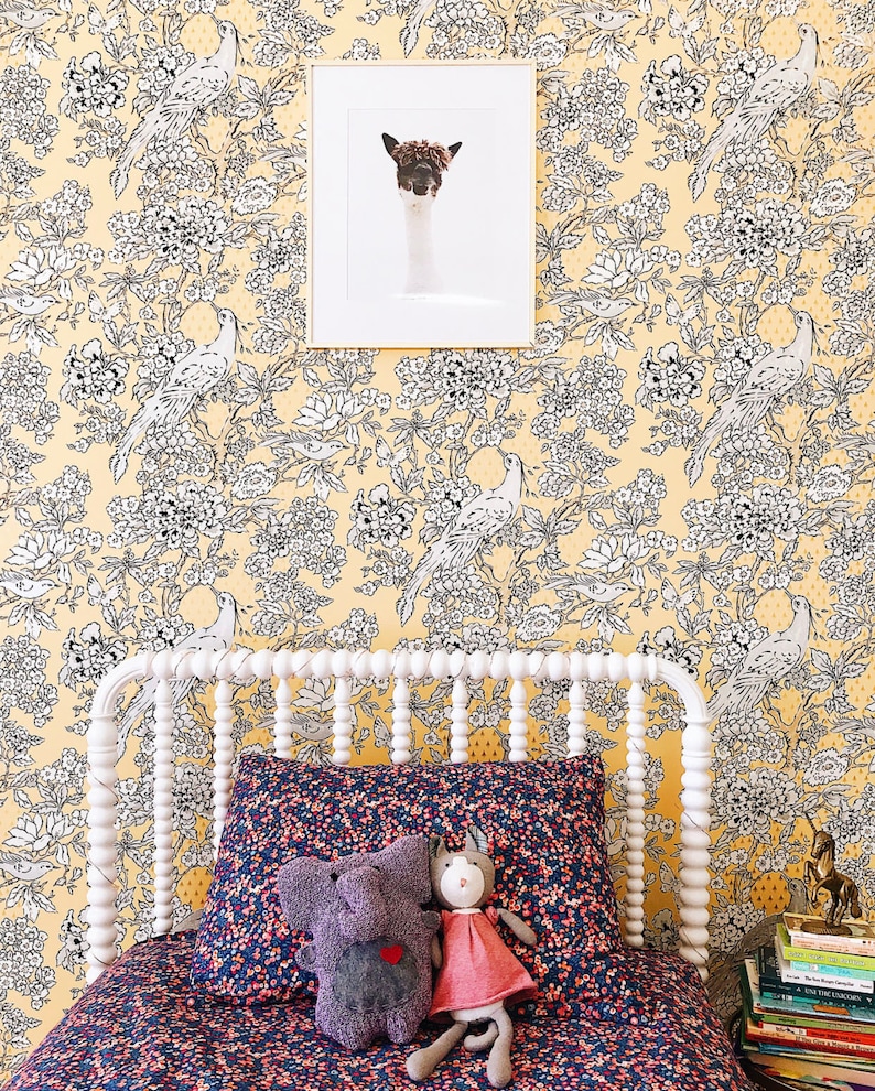Pu&ograve; includere: Un letto bianco con una testata e un piede a baldacchino bianchi. La testata ha un design decorativo di piccole perline bianche. Il letto &egrave; fatto con biancheria da letto a motivi floreali blu scuro e rosa. Due animali di peluche, un elefante grigio e un gatto rosa, sono seduti sul letto. La parete dietro il letto &egrave; rivestita di carta da parati gialla con un motivo floreale bianco e nero.