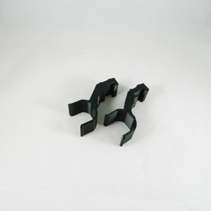 Könnte beinhalten: Zwei schwarze Kunststoffclips, jeweils mit einem gebogenen, offenen Design. Die Clips haben eine hakenartige Form an der Oberseite und sind vor einem weißen Hintergrund positioniert. Diese Clips werden wahrscheinlich zum Halten oder Sichern von Objekten verwendet.