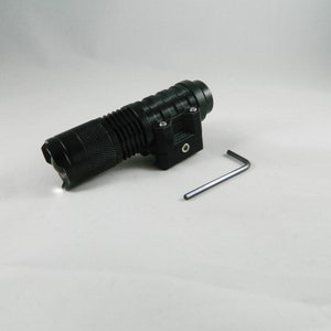 May include: Black flashlight with a textured handle and a mounting bracket. A small silver hex key is positioned next to the flashlight. The flashlight is angled towards the viewer, with the light illuminated.