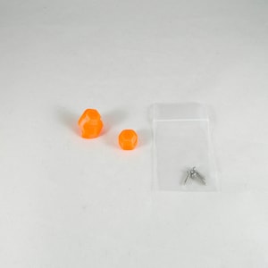 Op de afbeelding: Twee oranje plastic dobbelstenen en twee zilveren schroeven in een doorzichtige plastic zak.