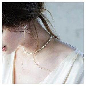 Puede incluir: En esta imagen se muestra un delicado collar de perlas y un pendiente largo y colgante. El collar está compuesto por pequeñas perlas redondas. El pendiente es una fina cadena dorada. La mujer lleva un vestido blanco.