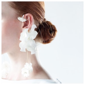以下が含まれることがあります： 白い花のイヤリングとイヤーカフのクローズアップ。イヤリングは、繊細な金色のワイヤーが付いた、大きくてフリルの花のデザインが特徴です。小さな花のイヤーカフも表示されます。背景は柔らかく、ニュートラルな白です。