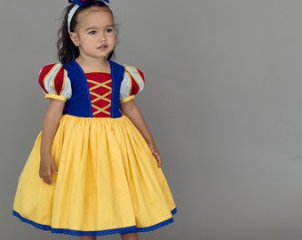 May include: A young girl dressed as Snow White in a yellow and blue dress with a red bow in her hair. She is standing on a gray background.