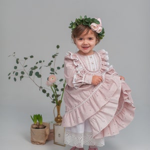 May include: A young girl wearing a pink floral dress with lace trim and a flower crown. She is standing in front of a white background with plants and a vase.