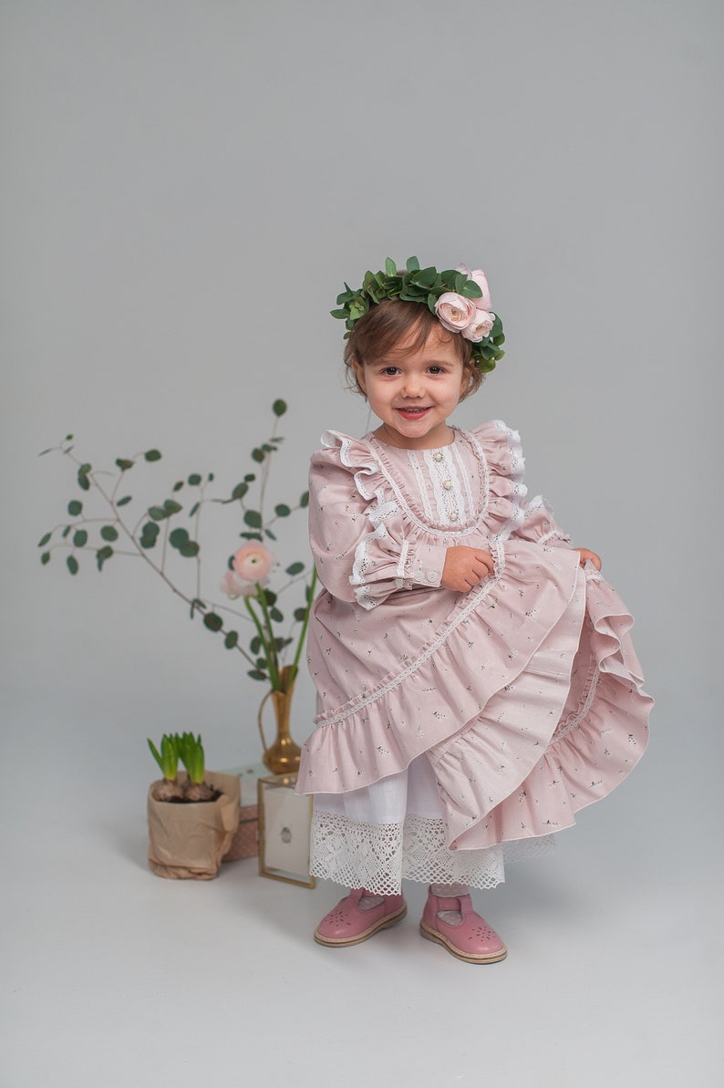 May include: A young girl wearing a pink floral dress with lace trim and a flower crown. She is standing in front of a white background with plants and a vase.
