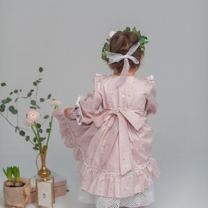 May include: A young girl wearing a pink floral dress with a white lace trim and a white ribbon in her hair. She is wearing pink shoes and has a wreath of green leaves on her head.