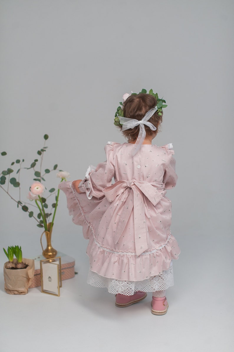 May include: A young girl wearing a pink floral dress with a white lace trim and a white ribbon in her hair. She is wearing pink shoes and has a wreath of green leaves on her head.