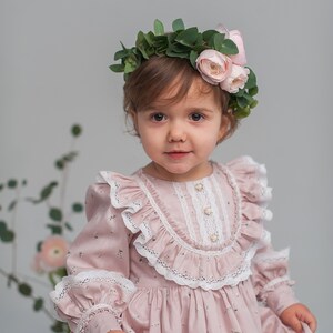 May include: A young girl wearing a pink dress with white lace trim and a floral crown. The dress has a floral pattern and the crown is made of green leaves and pink flowers.