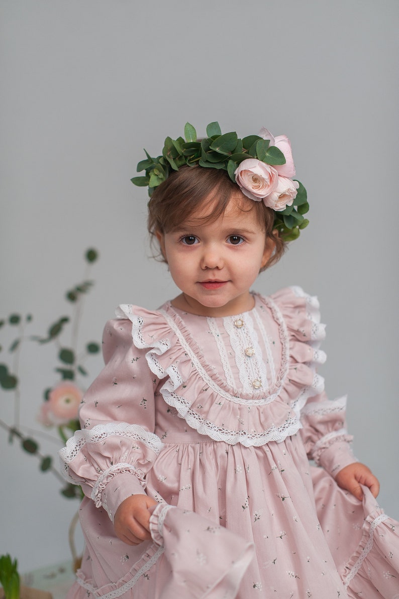 May include: A young girl wearing a pink dress with white lace trim and a floral crown. The dress has a floral pattern and the crown is made of green leaves and pink flowers.