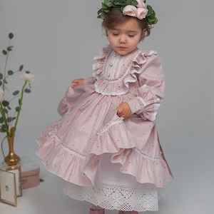 May include: A young girl wearing a pink floral dress with a white lace trim and a flower crown. She is wearing pink shoes and a white petticoat.