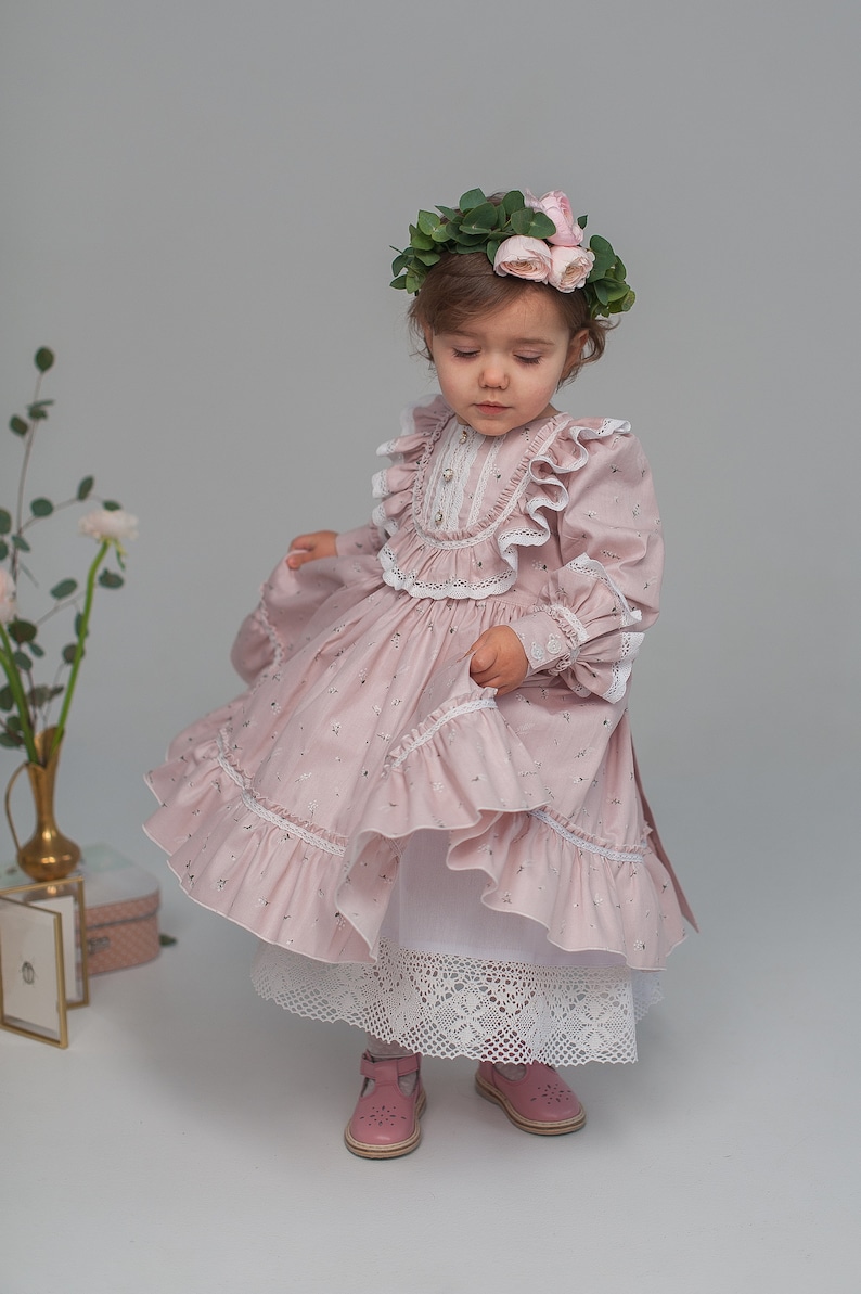 May include: A young girl wearing a pink floral dress with a white lace trim and a flower crown. She is wearing pink shoes and a white petticoat.