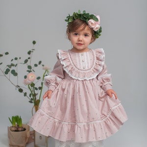 May include: A young girl wearing a pink floral dress with white lace trim and a flower crown. She is standing in front of a white background.