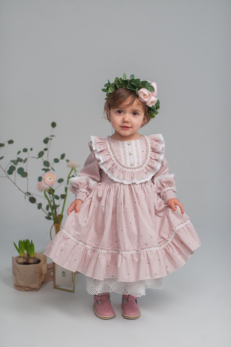 May include: A young girl wearing a pink floral dress with white lace trim and a flower crown. She is standing in front of a white background.