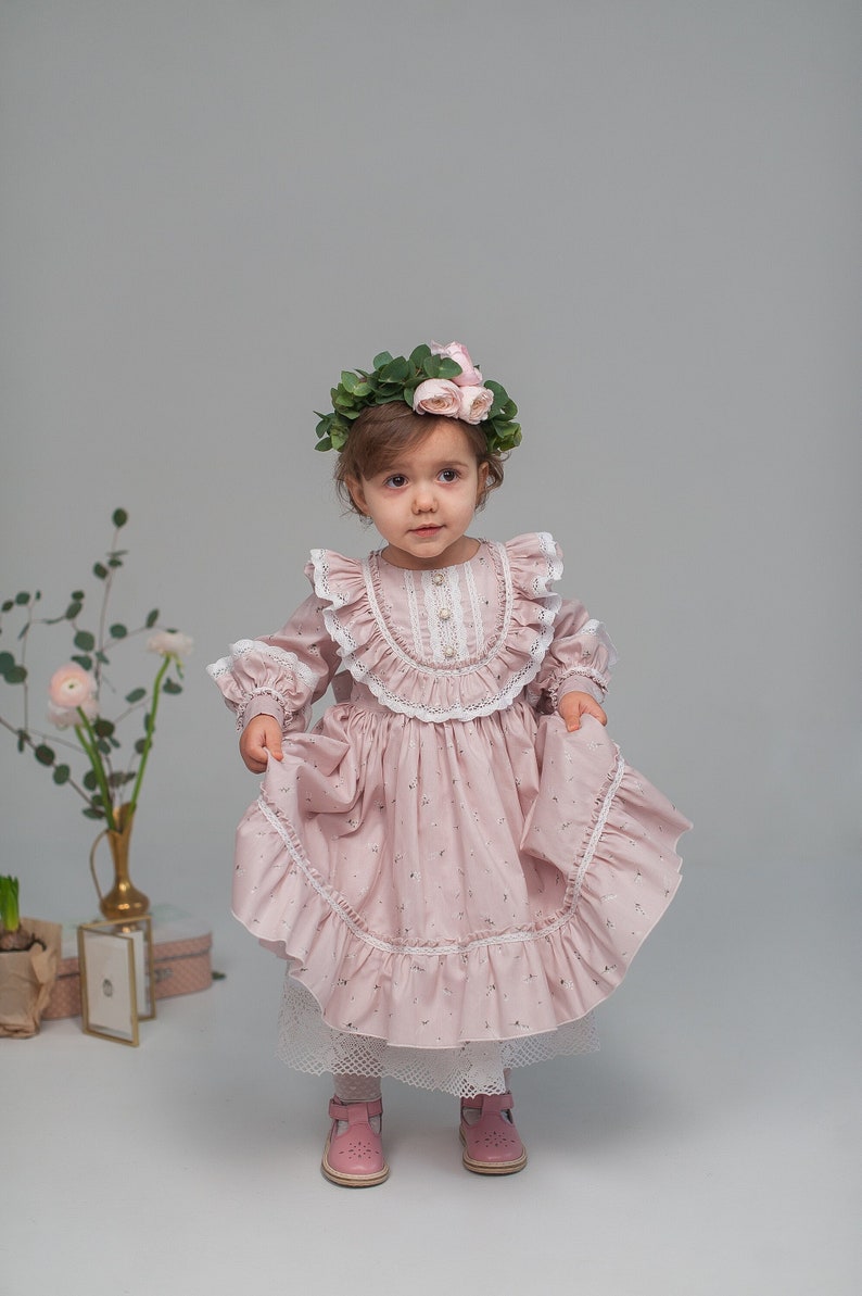 May include: A young girl wearing a pink floral dress with lace trim and a flower crown. She is standing on a white background.