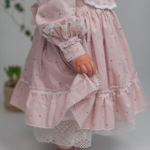 May include: A pink dress with a floral print and white lace trim. The dress has a ruffled collar and long sleeves with button cuffs. The dress is worn by a child and has a white petticoat underneath.