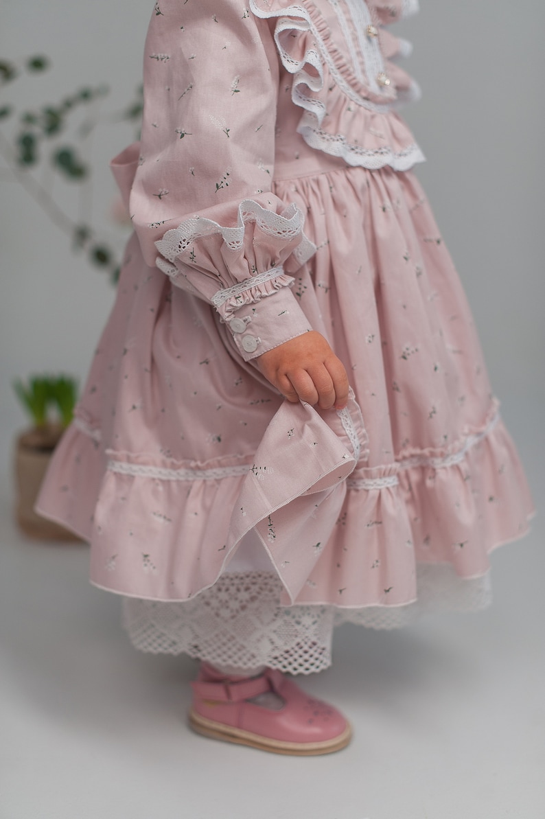 May include: A pink dress with a floral print and white lace trim. The dress has a ruffled collar and long sleeves with button cuffs. The dress is worn by a child and has a white petticoat underneath.