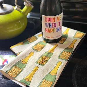 May include: A dark glass bottle with a label reading "OPEN ME WHEN IT HAPPENS Pop. Sip. Smile." sits on a white kitchen towel featuring a repeating pattern of green champagne bottles with text reading "FUCK TRUMP", "IS HE DEAD YET?", and "BIG BEAUTIFUL OBITUARY".