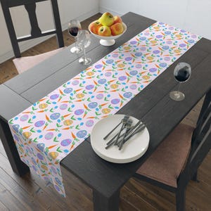 May include: A light pink Easter-themed table runner with a repeating pattern of decorated eggs, carrots, and blue tulips. The runner is placed on a dark wooden table, with a white bowl of apples and wine glasses.