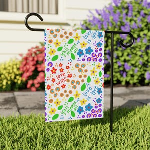 May include: A white garden flag featuring a colorful pattern of red, yellow, blue, and orange flowers, green leaves, and purple dots. The text reads "y'all means all", "love wins", and "love is love" in blue and red script across the surface.