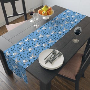 Pode incluir: Um caminho de mesa azul com um padrão repetido de pombas brancas e folhas verdes. O caminho de mesa está sobre uma mesa de madeira escura, com uma tigela branca de fruta e taças de vinho. A mesa tem cerca de 150 cm de comprimento.