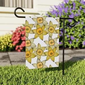May include: A garden flag featuring a repeating pattern of yellow daffodils with orange centers on a white background. The text "fuck mugs" is written on the center of each flower. The flag is displayed on a black metal stand in a garden setting.