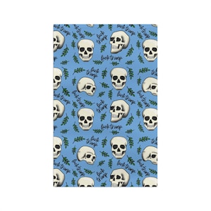 Puede incluir: Un textil azul con un patrón repetido de calaveras blancas, ramitas de hojas verdes y el texto manuscrito "fuck trump". Las calaveras están sombreadas y el diseño tiene un estilo gráfico audaz. El tema general es político.