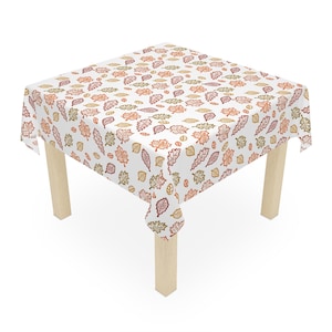 May include: A white tablecloth with a repeating pattern of autumn leaves in shades of orange, red, and green. The leaves are outlined in a detailed, artistic style. The tablecloth is draped over a square table with light-colored wooden legs.