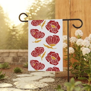 May include: A white garden flag featuring a repeating pattern of red flowers with gold stems and the word "HOME" in the center of each flower. The flag is displayed on a black metal stand in a garden setting.