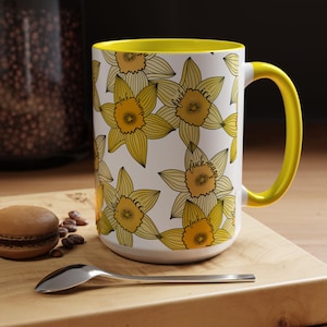 May include: White ceramic mug with a yellow interior and handle. The mug is decorated with yellow daffodil illustrations and the words "fuck ice" and "fuck maga". The mug is sitting on a wooden surface.